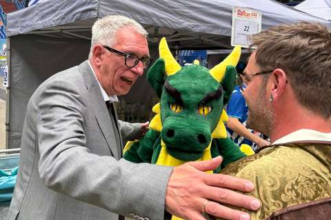 Drei Personen, darunter eine Person im grünen Drachenkostüm, stehen vor einem grauen Pavillon und unterhalten sich.