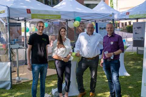 Vier Personen stehen nebeneinander vor weißen Pavillons mit Luftballons und Informationsständen im Freien.