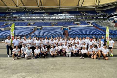 Eine große Gruppe von Personen in weißen T-Shirts steht und sitzt in einem Stadion vor leeren blauen Sitzreihen, flankiert von zwei grünen Fahnen.