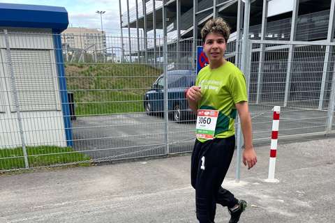 Eine Person mit Startnummer 3060 trägt ein grünes Laufshirt und schwarze Hose und geht auf einer Straße neben einem Zaun.