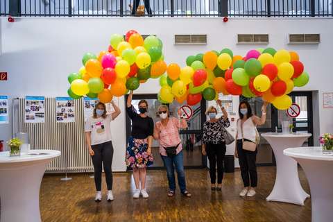 Fünf Personen mit Mund-Nasen-Schutz halten bunte Luftballons in einem Innenraum.