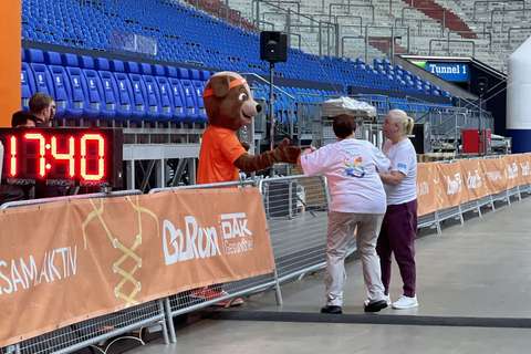 Zwei Personen geben einer Person im Bärenkostüm die Hand neben einer digitalen Uhr in einem Stadion.