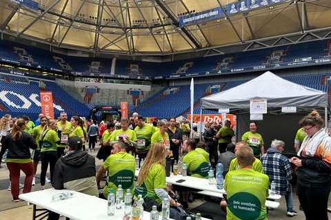 Mehrere Personen in grünen Laufshirts sitzen und stehen an Tischen in einem Stadion, im Hintergrund ein Zelt und leere Tribünen.