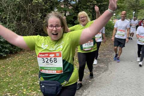 Mehrere Personen nehmen an einem Lauf im Freien teil, wobei eine Person im Vordergrund mit ausgebreiteten Armen in die Kamera läuft.