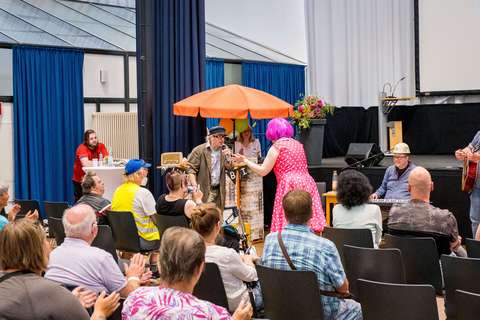 Mehrere Personen sitzen in einem Saal, während vorne eine Person mit pinker Perücke und gepunktetem Kleid sowie eine weitere Person mit Mikrofon auftreten; im Hintergrund steht ein orangefarbener Sonnenschirm.