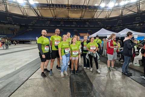 Eine Gruppe von acht Personen in grünen T-Shirts mit Startnummern steht in einer Halle, im Hintergrund befinden sich weitere Personen und weiße Zelte.