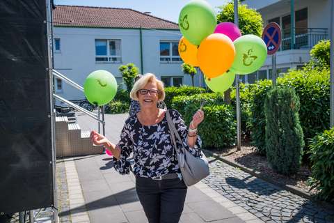 Eine Person mit Brille hält mehrere bunte Luftballons und trägt eine graue Tasche, im Hintergrund sind Gebäude und Grünpflanzen.