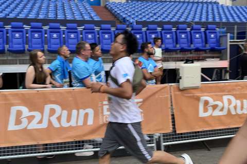 Eine Person läuft an einer Absperrung mit B2Run-Banner vorbei, im Hintergrund sitzen und stehen mehrere Personen auf blauen Stadionstühlen.