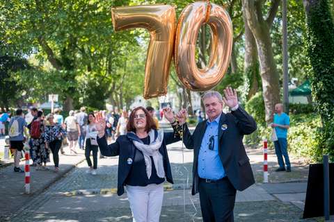 Zwei Personen halten goldene Luftballons in Form der Zahl 70 und winken, im Hintergrund sind weitere Menschen und Bäume.