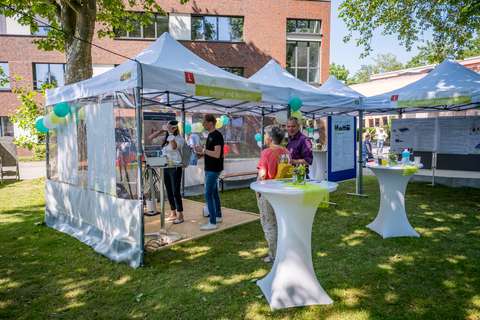 Mehrere Personen stehen und unterhalten sich an Stehtischen und unter Pavillons mit Luftballons, während eine Person eine Virtual-Reality-Brille trägt.