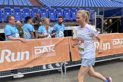 Eine Person läuft in Sportkleidung an einer Absperrung mit Werbebanner vorbei, im Hintergrund stehen mehrere Personen vor leeren Tribünen.