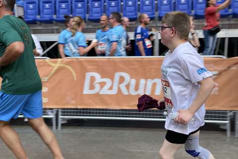 Eine Person mit weißem T-Shirt, kurzer schwarzer Hose und Kniebandage läuft in einem Stadion an einer Absperrung mit B2Run-Banner vorbei.