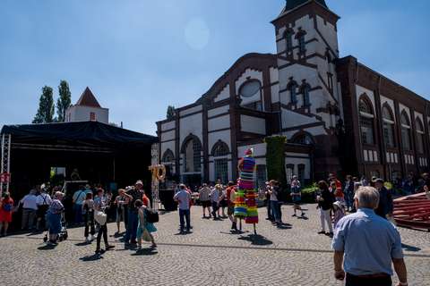 Mehrere Personen stehen und gehen auf einem gepflasterten Platz vor einem großen historischen Gebäude, eine Person auf Stelzen trägt ein buntes Kostüm, links steht eine Bühne mit der goldenen Zahl „70“ aus Luftballons.
