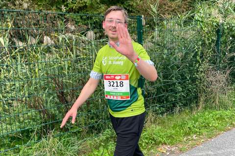 Eine Person mit Startnummer und grünem T-Shirt hebt eine Hand, geht an einem Zaun entlang und trägt eine Brille.