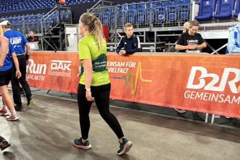Eine Person in Sportkleidung geht auf einer Halle an einer Absperrung entlang, im Hintergrund sitzen und stehen weitere Personen vor leeren Tribünen.