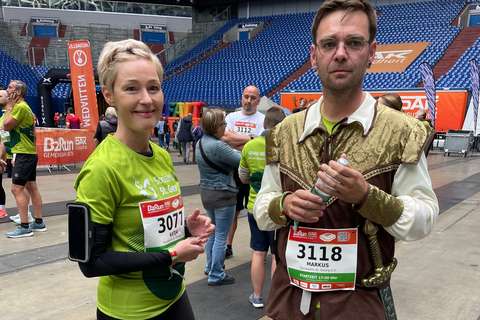Zwei Personen mit Startnummern stehen in einer Halle, eine Person trägt Sportkleidung, die andere ein historisches Kostüm und hält eine Wasserflasche.
