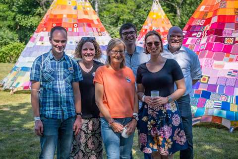 Sechs Personen stehen im Freien vor drei bunten Zelten aus Patchwork-Decken.