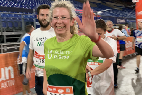 Eine Person mit Brille und grünem Laufshirt läuft und hebt eine Hand, weitere Personen laufen im Hintergrund.