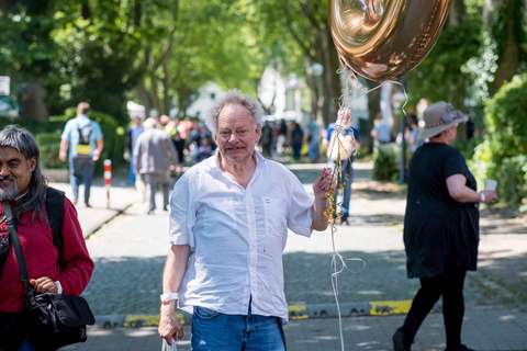 Eine Person mit weißem Hemd hält einen goldenen Ballon und eine Tüte, im Hintergrund gehen weitere Personen auf einer baumbestandenen Straße.