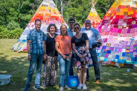Sechs Personen stehen auf einer Wiese vor bunt gestrickten Tipis.
