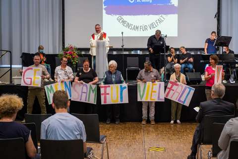 Mehrere Personen stehen nebeneinander und halten bunte Schilder mit den Buchstaben „GEMEINSAM FÜR VIELFALT“.