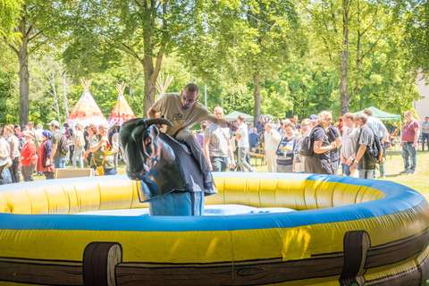 Eine Person reitet auf einem mechanischen Bullen in einer gelben Hüpfburg, im Hintergrund stehen viele Personen und zwei bunte Tipis unter Bäumen.