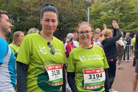 Zwei Personen mit grünen Laufshirts und Startnummern stehen nebeneinander in einer Gruppe von Menschen im Freien.