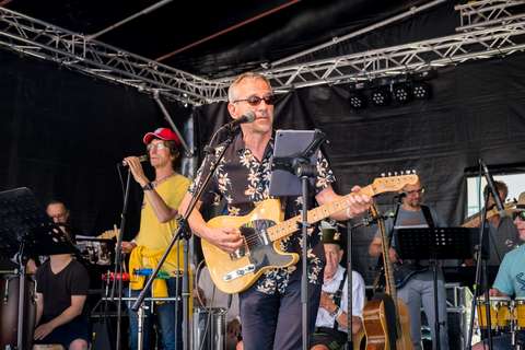 Mehrere Personen musizieren auf einer Bühne, eine Person im Vordergrund spielt E-Gitarre und singt in ein Mikrofon.