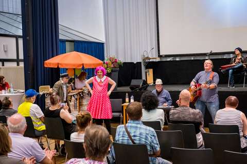 Mehrere Personen führen auf einer Bühne und davor Musik auf, eine Person trägt ein pinkes Kleid mit Punkten und eine pinke Perücke, im Hintergrund sitzen Menschen im Publikum.