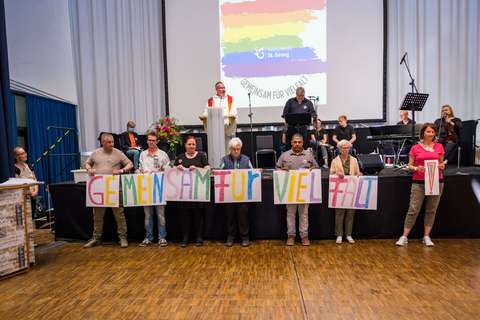 Mehrere Personen stehen nebeneinander und halten bunte Schilder mit der Aufschrift „GEMEINSAM FÜR VIELFALT“ vor einer Bühne, auf der weitere Personen sowie Musikinstrumente zu sehen sind.