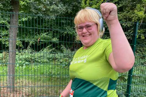 Eine Person mit Kopfhörern und grünem T-Shirt hebt den Arm und lächelt, im Hintergrund ein Zaun und Bäume.