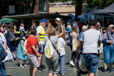 Mehrere Personen stehen und gehen auf einem Platz im Freien, einige tragen Stoffbeutel auf dem Rücken, eine Person hebt die Arme, eine Person schiebt einen Rollstuhl.
