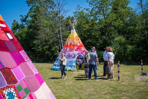 Mehrere Personen stehen auf einer Wiese vor einem Tipi, das mit bunten Patchwork-Decken bedeckt ist; im Vordergrund ist ein weiteres buntes Patchwork-Objekt zu sehen.
