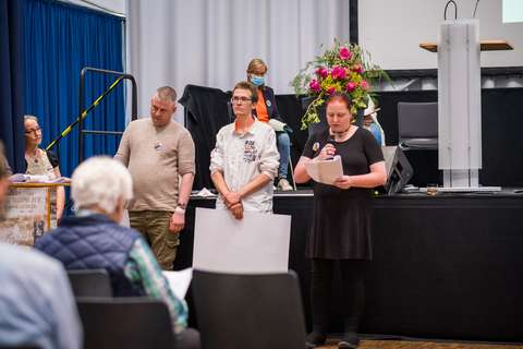 Drei Personen stehen nebeneinander, eine Person hält ein Mikrofon und liest von einem Blatt Papier, im Hintergrund sitzt eine weitere Person vor einem Blumenarrangement.