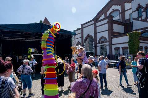 Mehrere Personen stehen und gehen auf einem Platz vor einem Gebäude, eine Person auf Stelzen trägt ein buntes Kostüm aus Ringen, im Hintergrund ist eine Bühne aufgebaut.