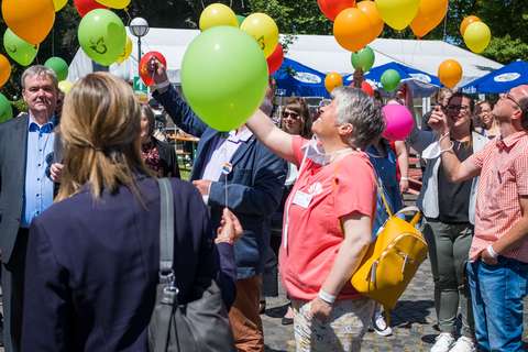 Mehrere Personen stehen im Freien auf Kopfsteinpflaster und halten bunte Luftballons.