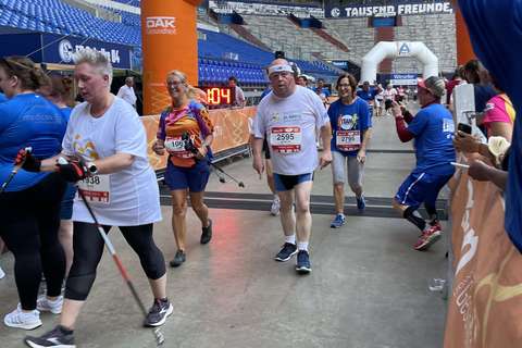 Mehrere Personen überqueren mit Startnummern und Sportkleidung eine Ziellinie unter einem orangefarbenen Torbogen in einem Stadion.
