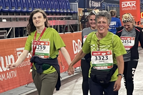 Zwei Personen mit Startnummern und grünen T-Shirts gehen Hand in Hand auf einer Laufveranstaltung.
