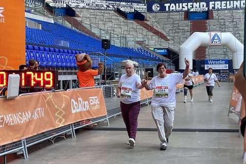 Zwei Personen laufen gemeinsam unter einem orangefarbenen Zielbogen bei einer Laufveranstaltung in einem Stadion, im Hintergrund steht ein Maskottchen.