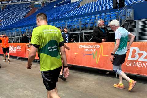 Mehrere Personen in Sportkleidung gehen an einer Absperrung mit orangefarbenem Banner entlang, im Hintergrund leere blaue Tribünen.