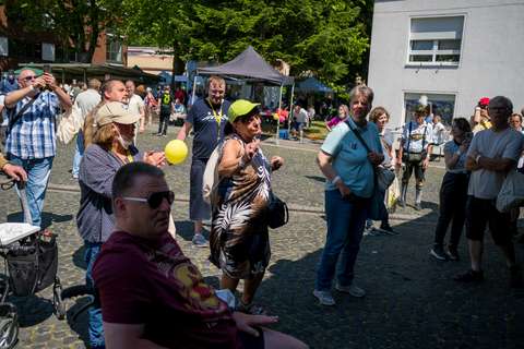 Mehrere Personen stehen und sitzen auf einem gepflasterten Platz im Freien, einige halten Ballons oder Handys, im Hintergrund sind Marktstände und Bäume.