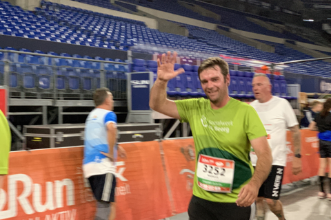 Eine Person mit grünem T-Shirt und Startnummer winkt, während sie an einer Absperrung in einem Stadion vorbeiläuft.
