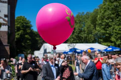 Ein pinker Luftballon schwebt im Vordergrund, im Hintergrund stehen mehrere Personen im Freien vor weißen Zelten und Bäumen.