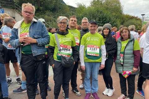 Sieben Personen stehen in Sportkleidung und mit Startnummern nebeneinander im Freien, umgeben von weiteren Menschen.
