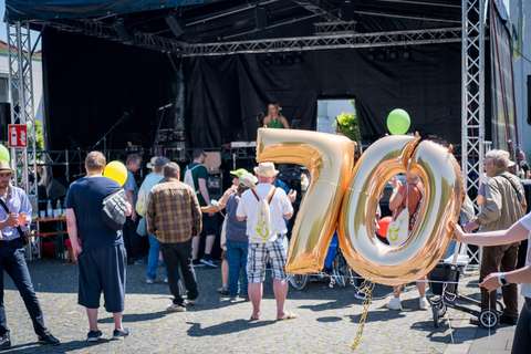 Mehrere Personen stehen vor einer Bühne im Freien, eine Person hält goldene Luftballons in Form der Zahl 70.