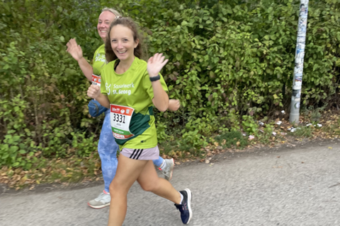 Zwei Personen joggen auf einer Straße und winken, beide tragen grüne T-Shirts mit Startnummern.