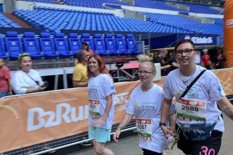 Drei Personen mit Startnummern und weißen T-Shirts gehen nebeneinander in einem Stadion.