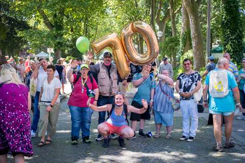 Eine Gruppe von Personen steht im Freien, einige halten goldene Luftballons in Form der Zahl 70 und einen grünen Ballon, eine Person hockt vorne.