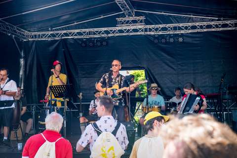 Mehrere Personen musizieren auf einer Bühne, während im Vordergrund weitere Personen stehen und zuhören.