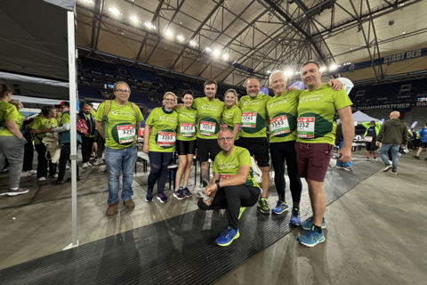 Eine Gruppe von neun Personen in grünen Sportshirts mit Startnummern steht und hockt in einer Halle.